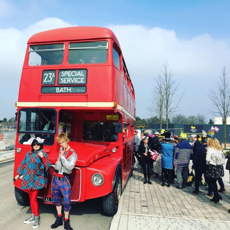 A man and woman in fashionable clothes stand in front of a red double decker bus as passengers queue up to get on