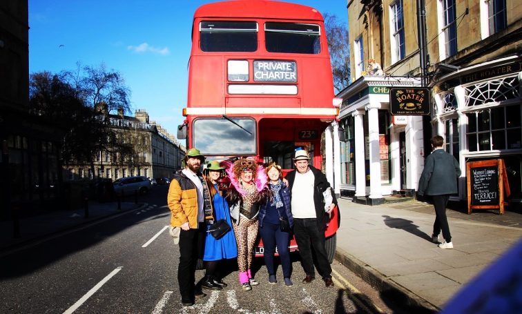 A vibrant group of people stand in front of an old fashioned red double decker bus