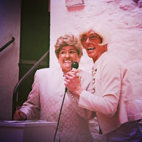 2 men in male drag hold a microphone and look towards an audience as they host a fashion show
